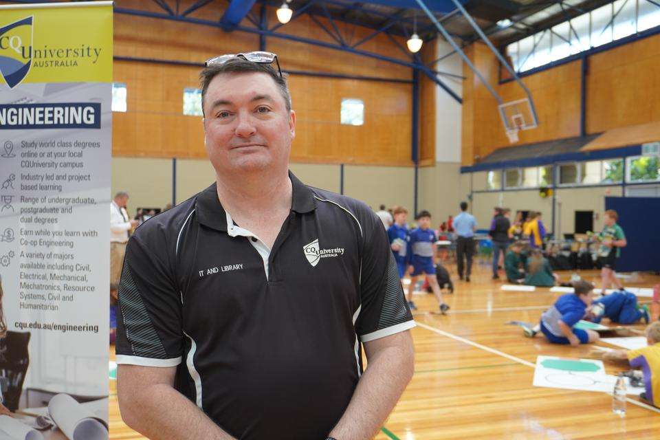 CQU Robotics Comp Organiser and IT staff Jason Bell stands in a sports stadium with a CQU Engineering banner. Students are engaged in various activity in the background.