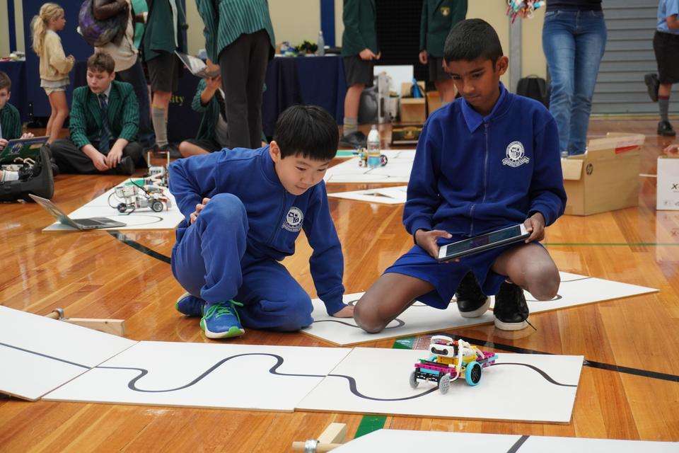 Two primary students sit on the floor of a sports stadium with their mechanised robot, iPad controller and competition mat. Students are pictured in various activity behind them.
