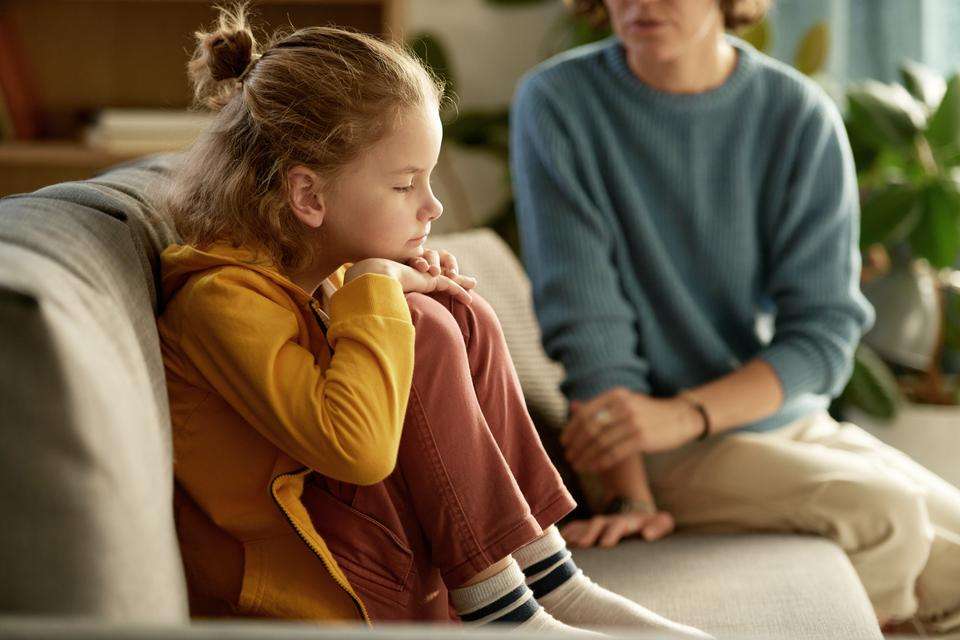 Young child sits side on, on a couch clutching their knees and looking sad. They are not looking into the camera. A woman, perhaps their mother, sits in the background and appears to be trying to reassure the child.