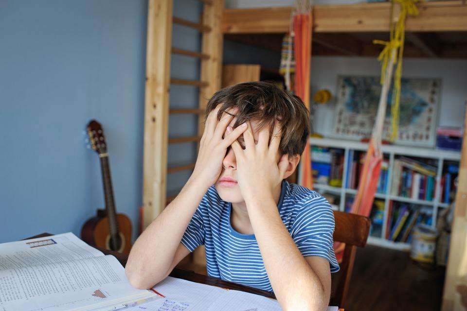 Young boy sits at a desk with head in hands with homework in front of him. There is a bookcase full of books and guitar in the background.