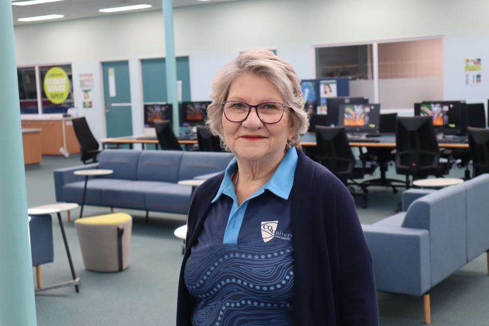 Louise Geal stands in a library in front of a bank of computers, she wears a blue shirt with CQU logo and Indigenous pattern.