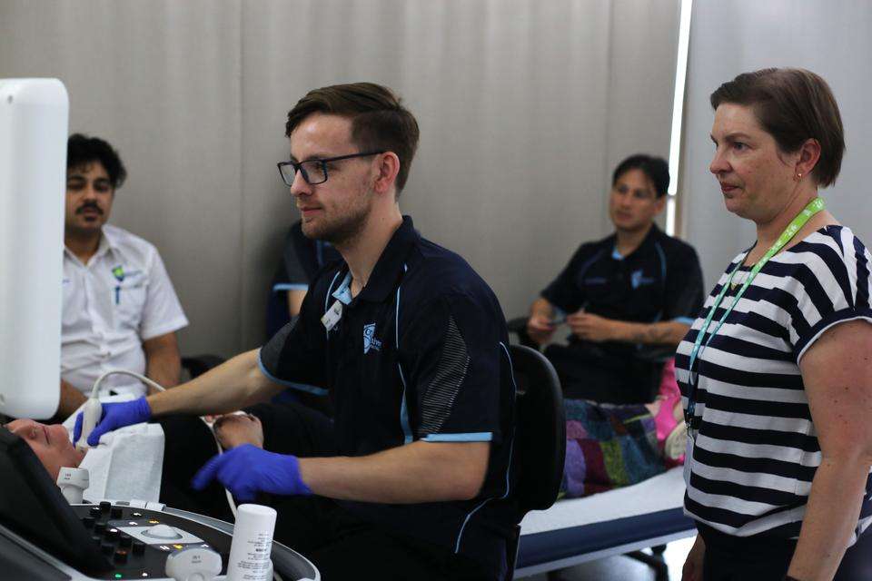 A sonography student uses ultrasound equipment to scan a patient, while teacher and students watch.