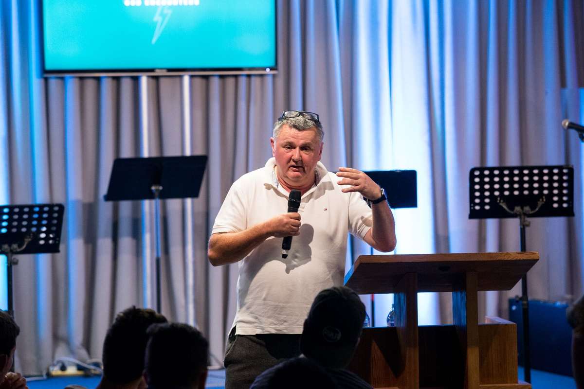 A man in a white polo shirt holding a microphone on a stage with an audience in the foreground