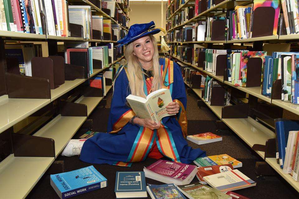 Dr Shirley Ledger sits on floor in library, surrounded by social work books, wearing doctoral regalia.