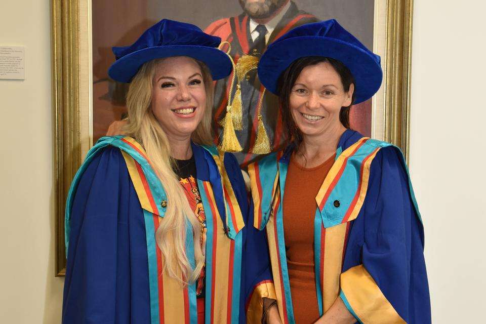 Dr Shirley Ledger and Prof Susan Kinnear stand in front of a portrait of a former vice-chancellor. They both wear colourful doctoral regalia.