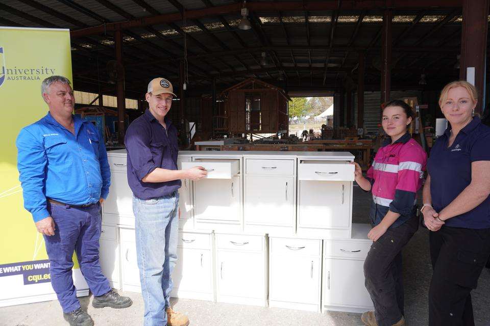 A CQU teacher, two students, and a stakeholder from Roseberry QLD stand in a trades workship with 11 bedside tables and a CQUniversity pull-up banner. The students are holding the cabinet drawers open in demonstration