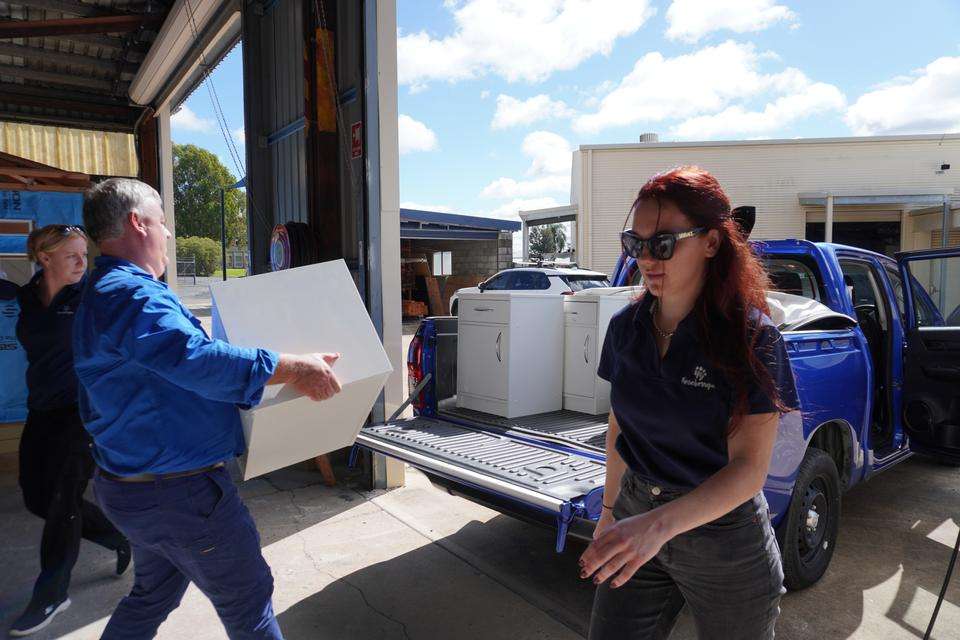 A man loads a bedside table onto a ute tray as two women walk by to help