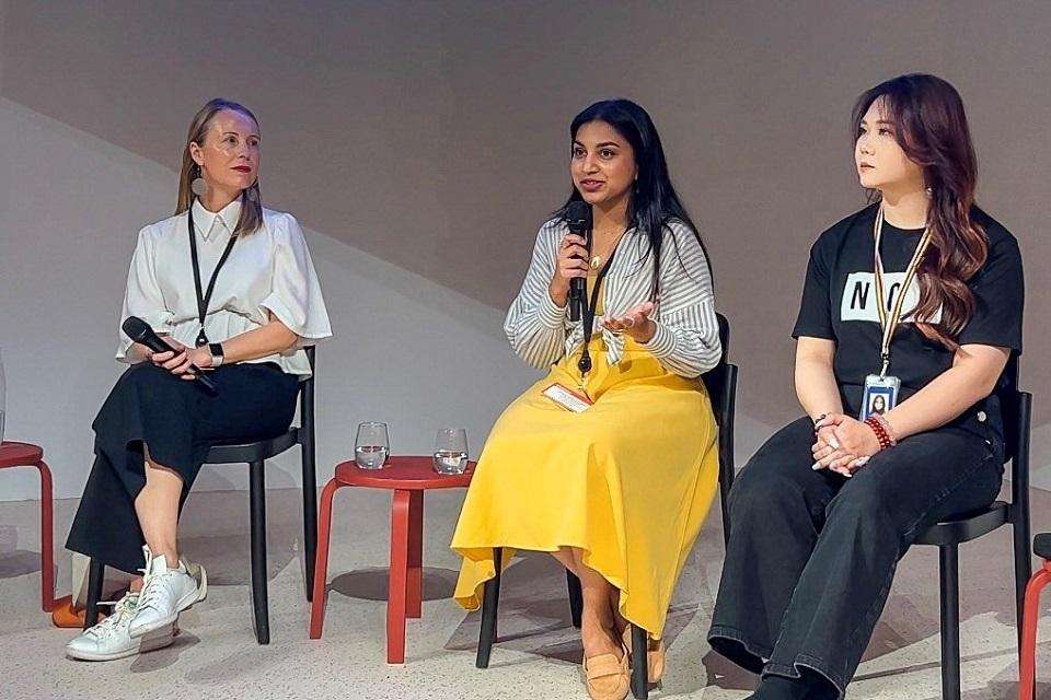 CQU student Amanda Abeysinghe (centre) wears yellow skirt and striped top, sits between two other women, holds a microphone.