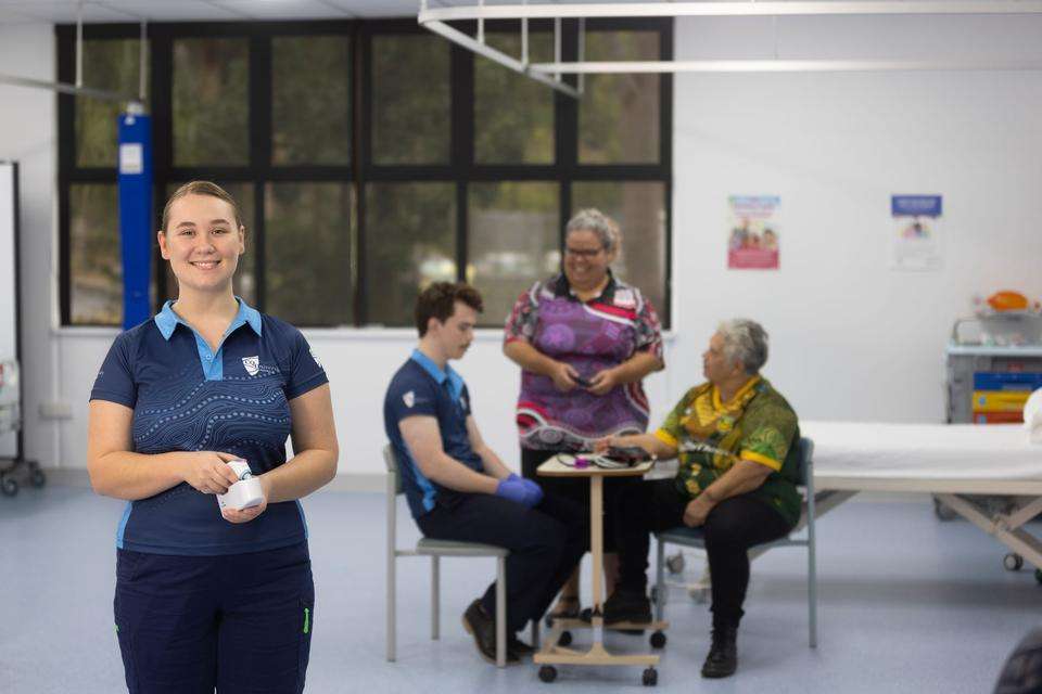 A young woman stands in the foreground of a clinical setting holing a thermometer. Three others are pictured in the background taking someone's blood pressure.