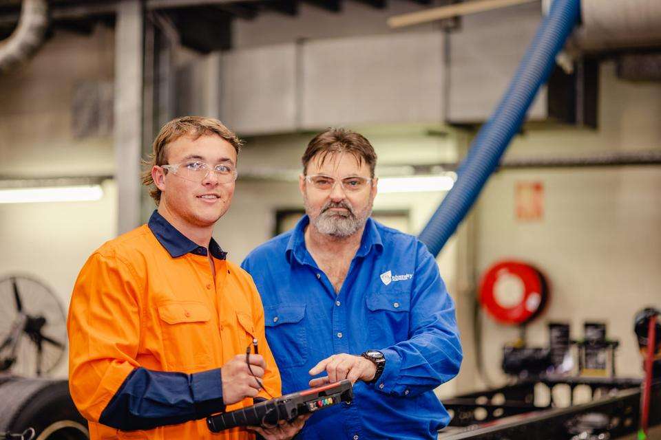 Two men are pictured in a workshop setting. Both wear PPE. One is holding a technical tool used in automotive diagnostics.