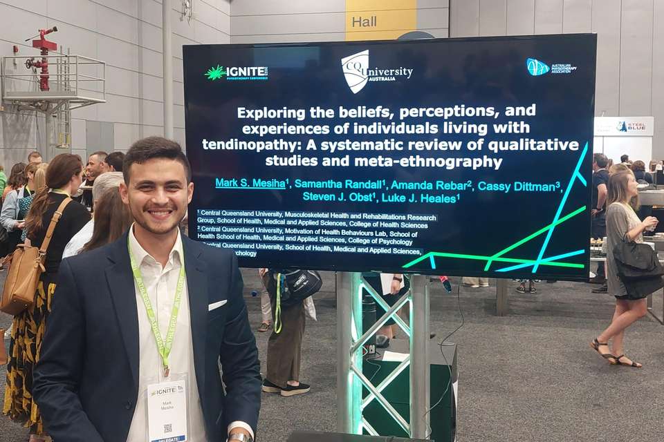 Mark Mesiha stands in the hall of a busy convention centre; people mill about in the background and he stands smiling, in front of a digital screen that displays the title of his research paper on tendinopathy.