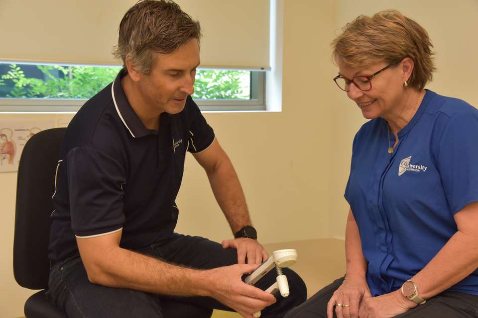 Dr Luke Heales sits with a model patient in a clinical examination room; the two people look at a clinic instrument together to read the results.