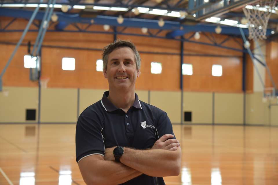 Dr Luke Heales stands in a sports/basketball stadium, smiling with his arms crossed