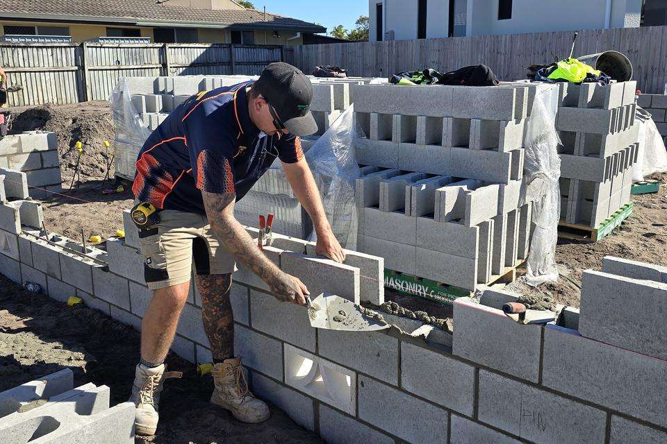 A young man wearing a cap and holding a trowel to a brick wall