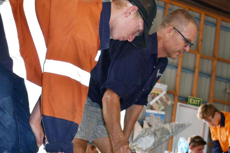 A young man in a hi-vis shirt receives instruction from a man with short hair in a blue shirt and grey pants holding a trowel. In the background are some other young men using trowels.