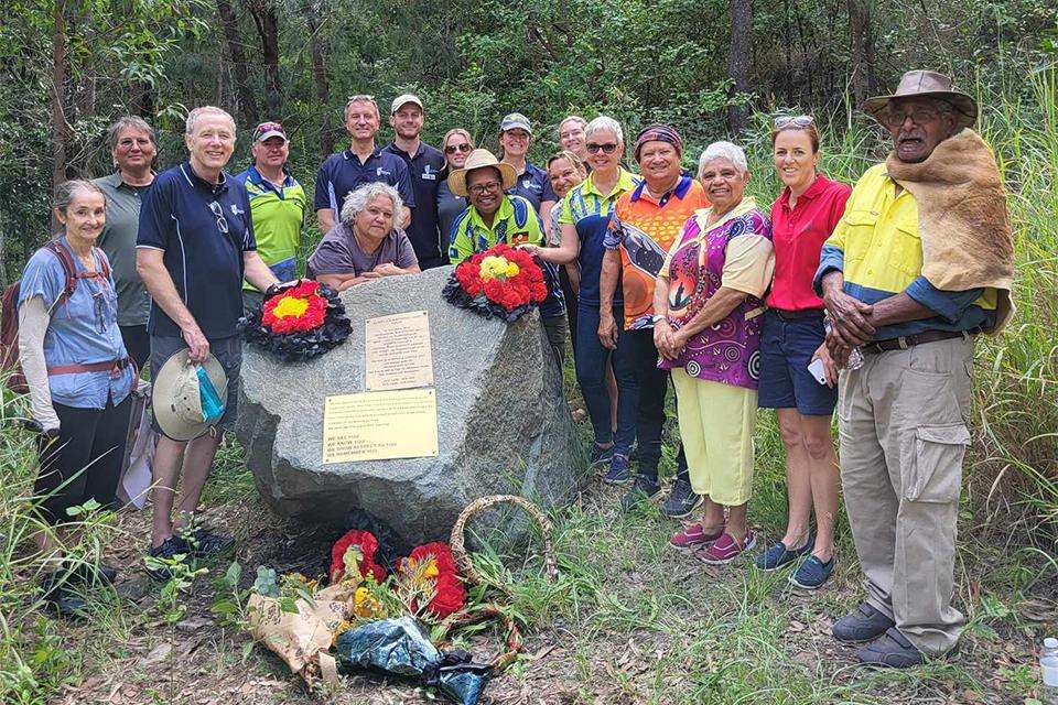 CQUniversity staff, students and alumni with Darumbal Elders at Gai-i for an an Honouring Ceremony stand in front of a commemorative memorial with wreaths in the colours of the Aboriginal Flag