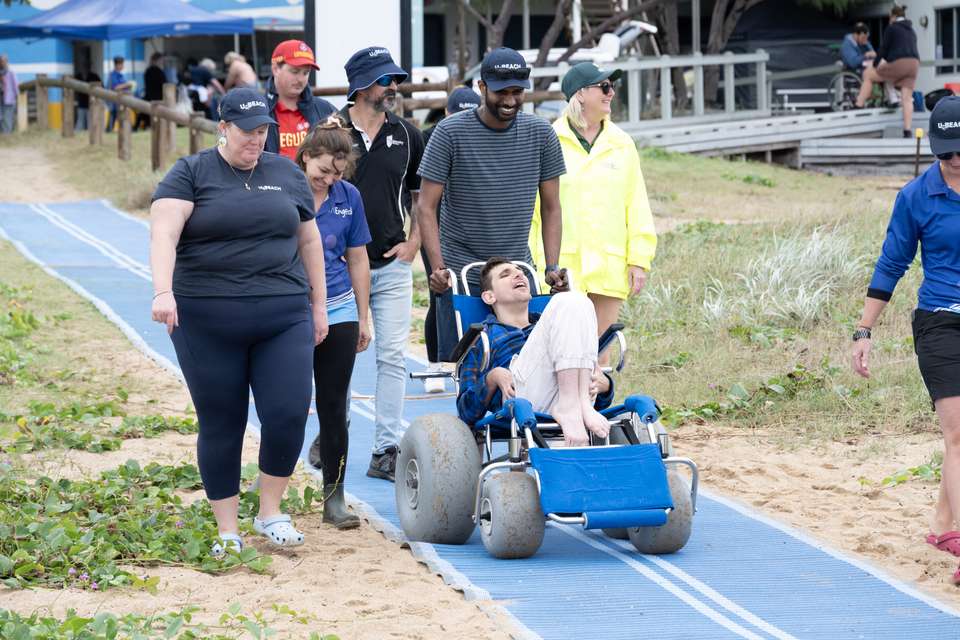 A group of people assisting a man in a wheelchair use a beach mat