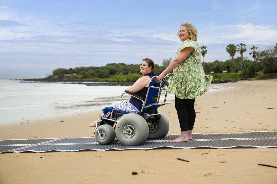 A student helping push another student in a wheelchair on the beach looking out at the ocean smiling.