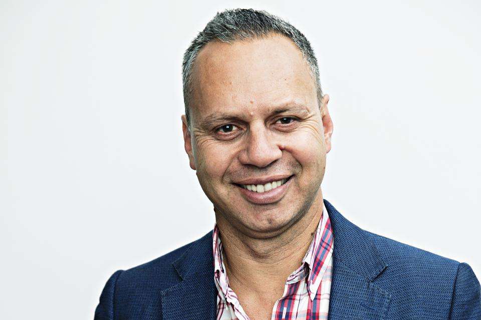 Man with short hair looking at camera with a broad smile, wearing red and blue chequered business shirt and a navy blue jacket