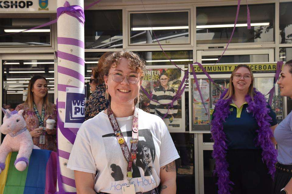 CQU staff member and Ally Program member Gabby Rothery stands smiling at the camera with people in the background smiling and chatting with each other as they celebrate Wear It Purple Day