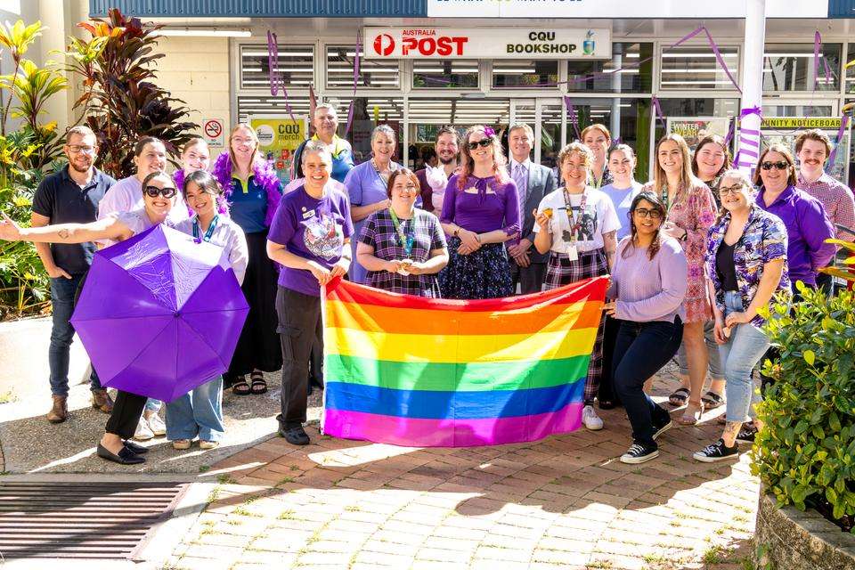 A group of people pose for a photo standing outside on a sunny day, wearing purple and holding a rainbow flag to celebrate Wear it Purple Day