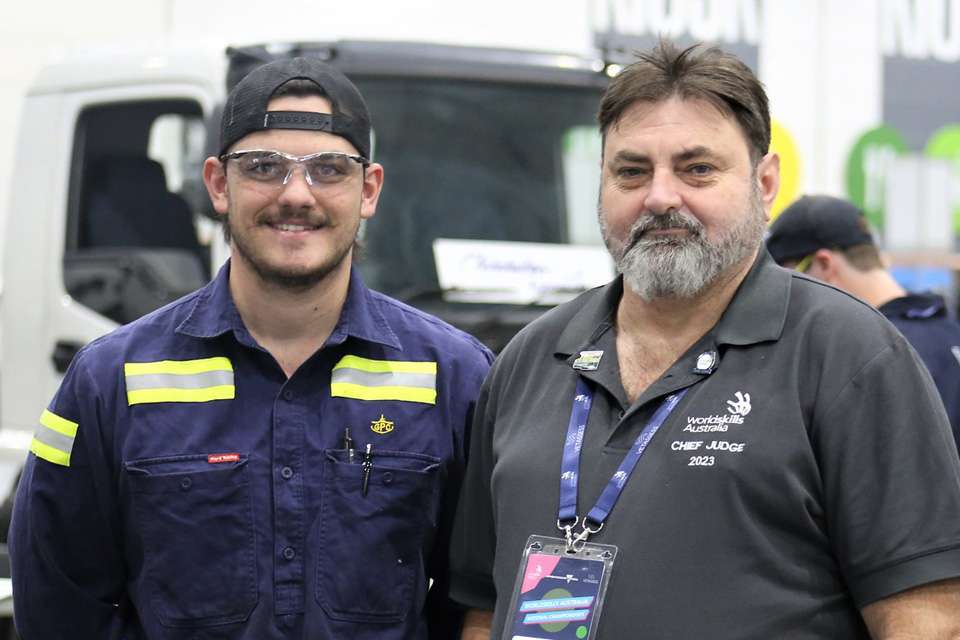 Two men standing side by side, one wearing a navy hi-viz shirt and backwards baseball cap and protective goggles the other has a gray beard and is wearing a gray shirt with the words worldskills australia chief judge on it