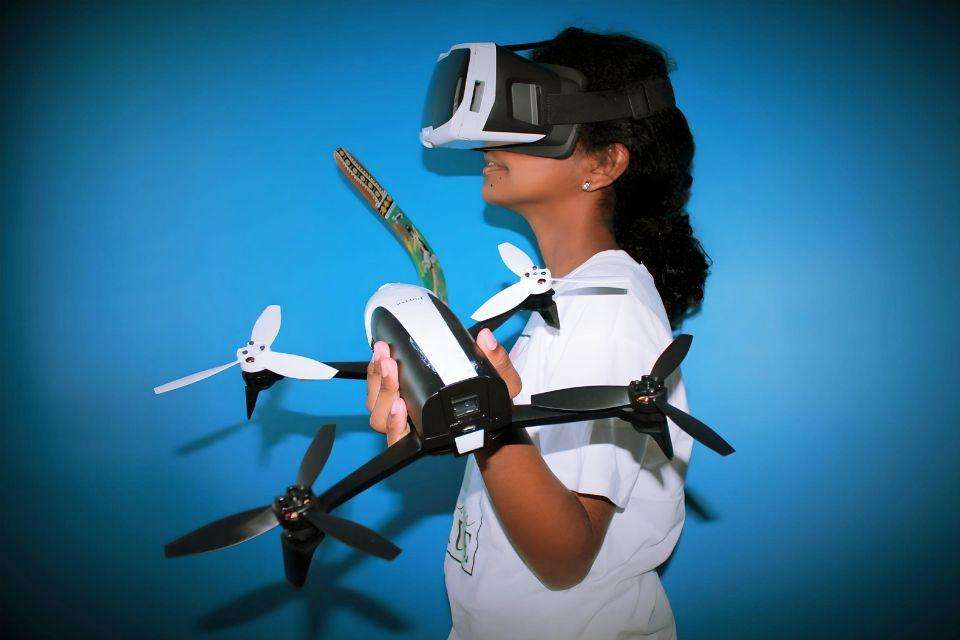 A girl with black hair wears a virtual reality headset, and holds a drone and a boomerang, standing in front of a blue wall.