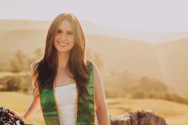 Nicole Shanley smiling, wearing a Cal Poly graduation sash, leaning against a rock with fields in the background