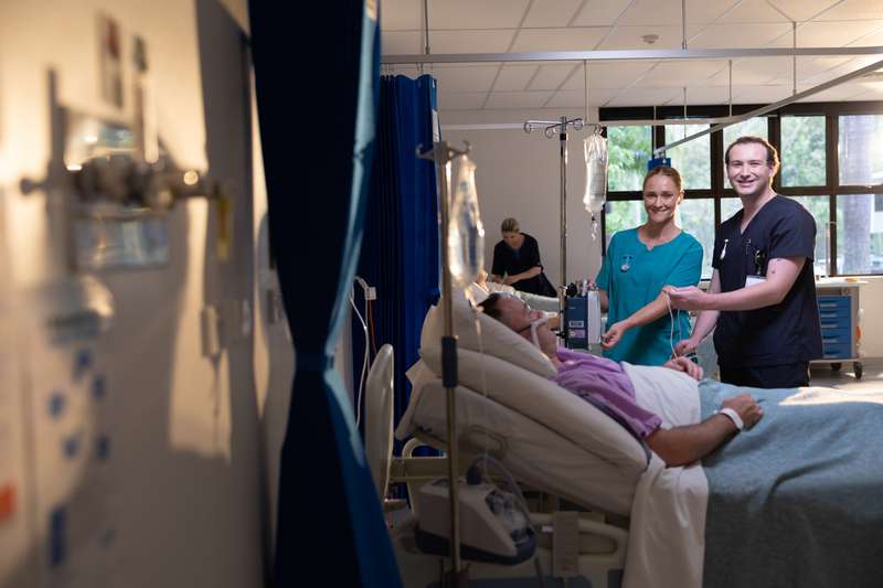 Two nursing students caring for a patient who is in bed in a realistic hospital setting.