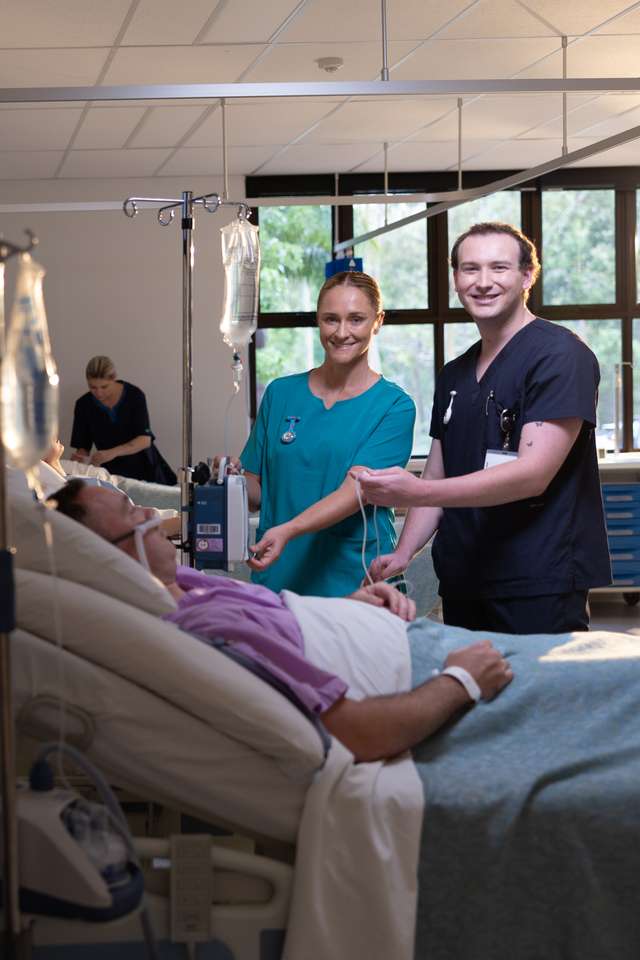 Two nursing students caring for a patient who is in bed in a realistic hospital setting.