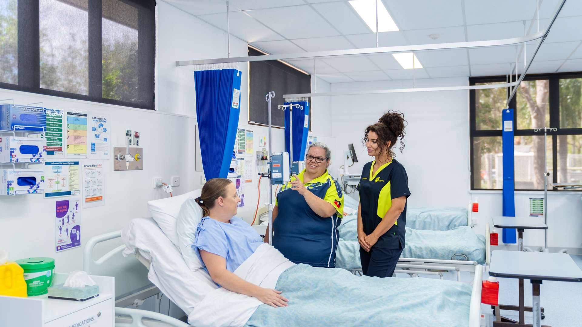 A nursing lecturer assists a nursing student in caring for a patient who is in bed in a realistic hospital setting.