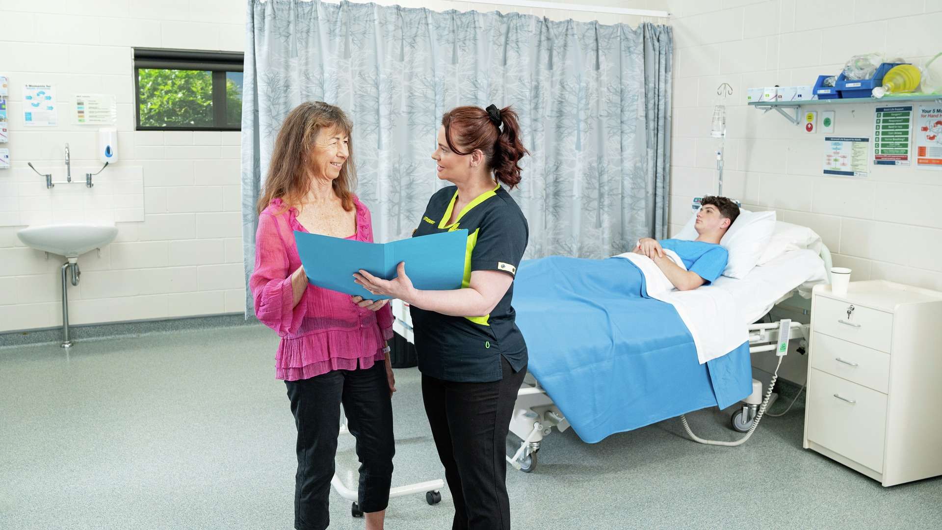 A nursing student and teacher discussing a patient's file with patient lying in hospital bed in background