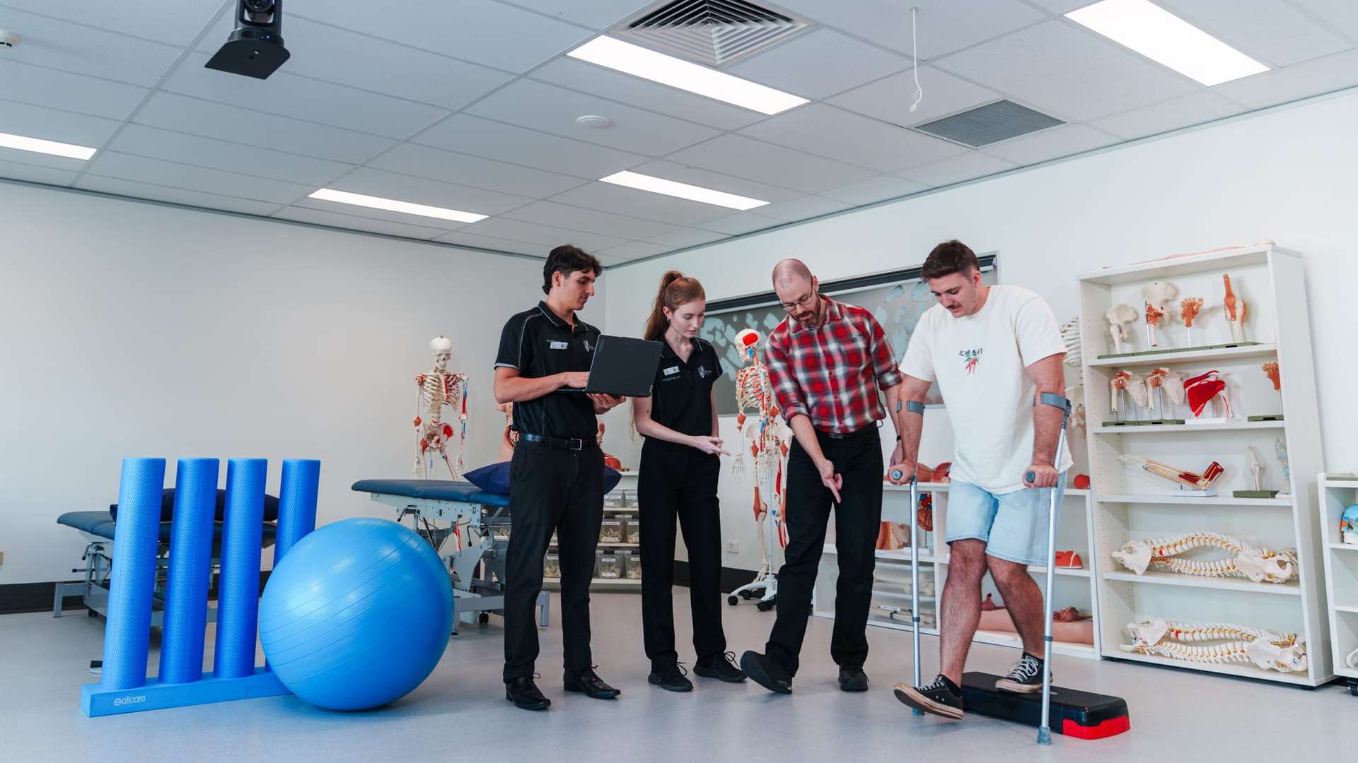 A group of people in a physiotherapy training room observe and assist someone using a balance step.