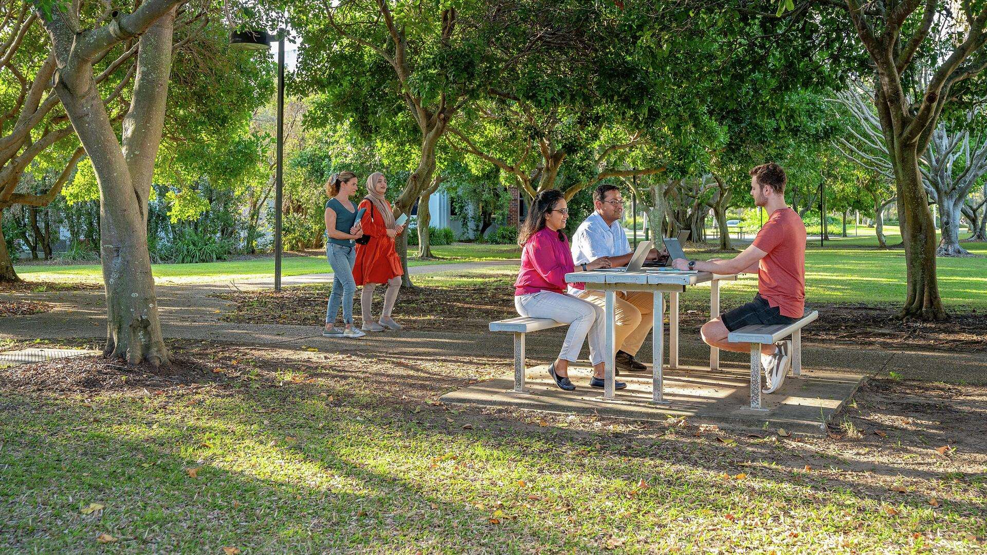 Three students studying outside at table on CQU Gladstone campus with two students walking on path