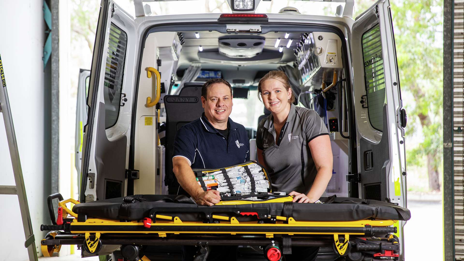CQUniversity Paramedics standing in the back of an open ambulance, smiling and holding a medical kit on a stretcher.