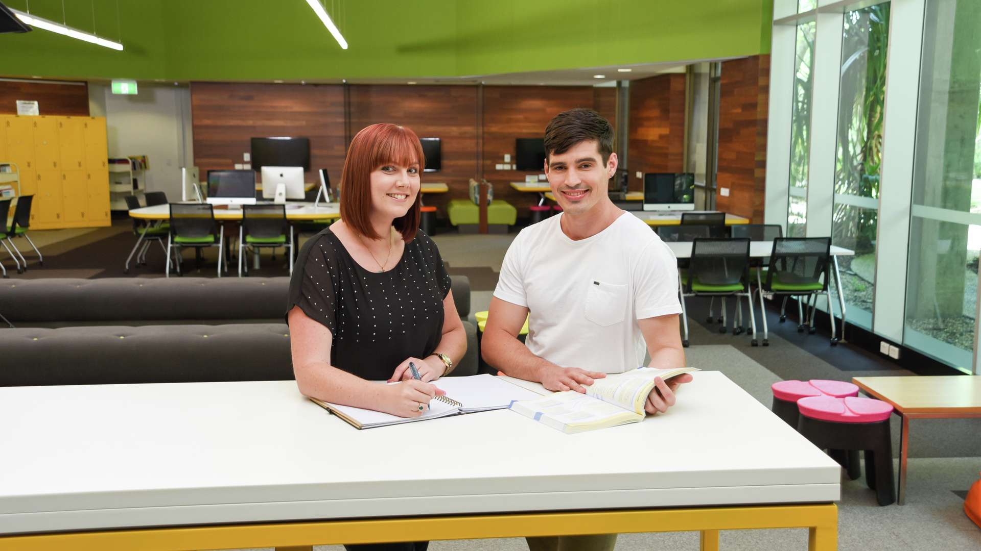 Two students standing at table in CQU Rockhampton library with text book and notepad.