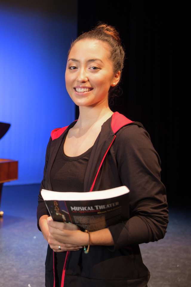 Musical theatre student holding a textbook on stage with lecturer playing piano in background