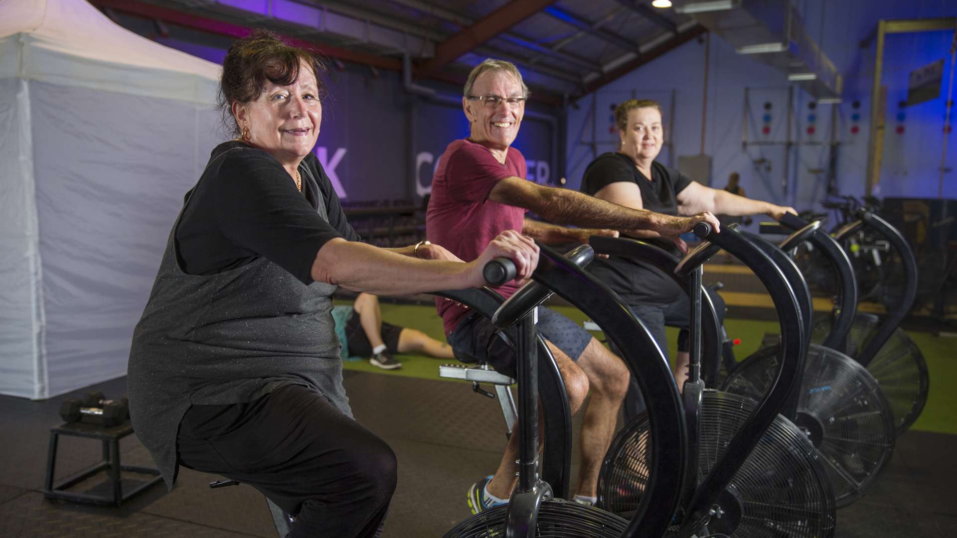 Three people are seated on exercise bikes in an indoor gym, exercising side by side.