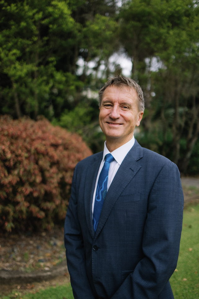 Nick Klomp wearing a navy suit, standing in front of greenery and smiling at camera with arms at his side