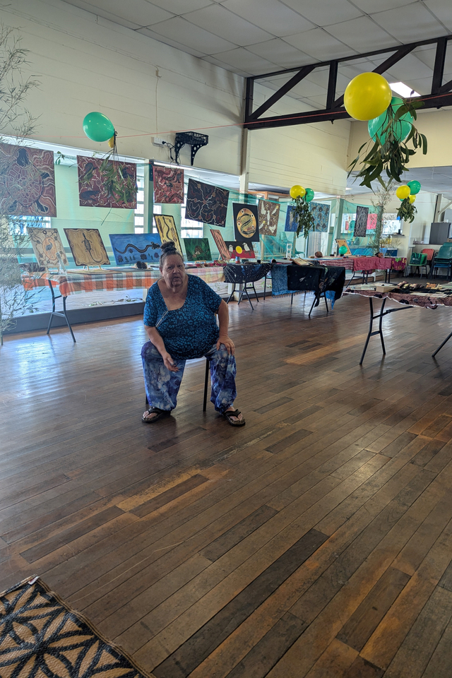 A person sits on a chair in a large hall with wooden floors and tables display artworks.