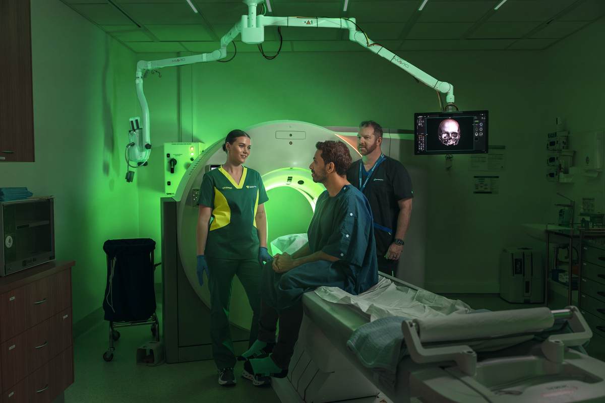 Student talking to a paitent who is seated on a MRI machine as a Queensland X-Ray staff memebr watches on