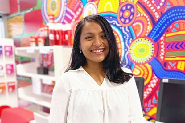 Rajani Shrestha, from Nepal, smiling in front of a decorative Indigenous Australian art wall mural, and desktop computers