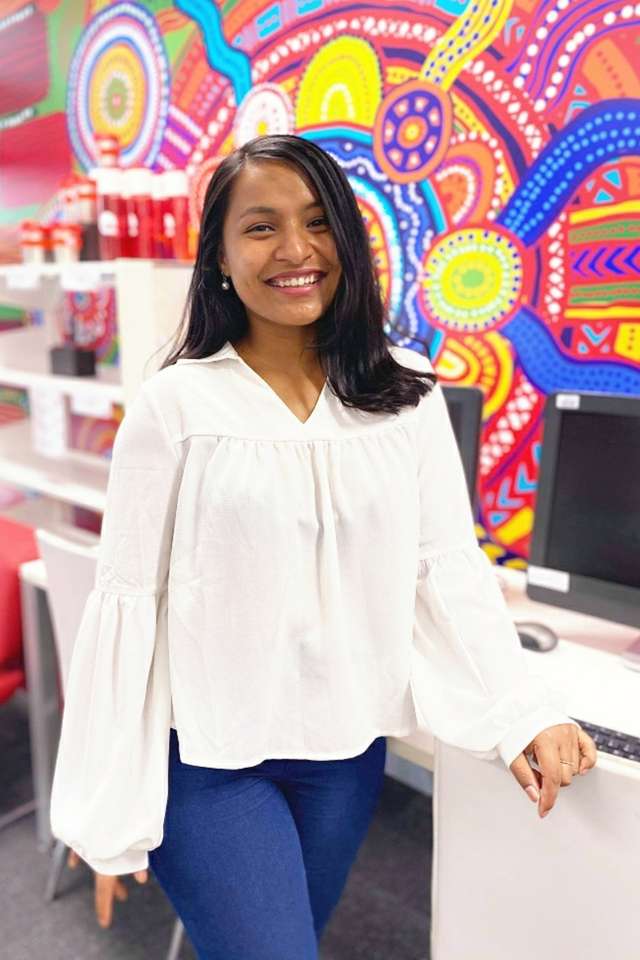 Rajani Shrestha, from Nepal, smiling in front of a decorative Indigenous Australian art wall mural, and desktop computers