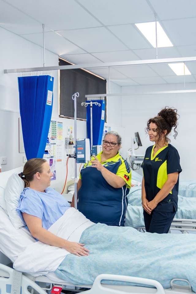 A nursing lecturer assists a nursing student in caring for a patient who is in bed in a realistic hospital setting.
