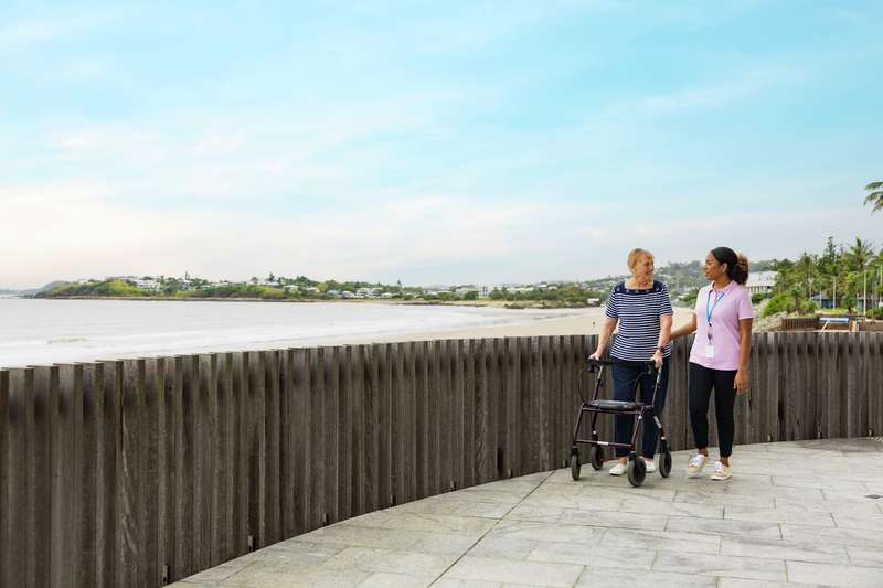 A CQU Community Services student takes a stroll along the esplanade at the beach with an elderly patient who requires assistance with walking