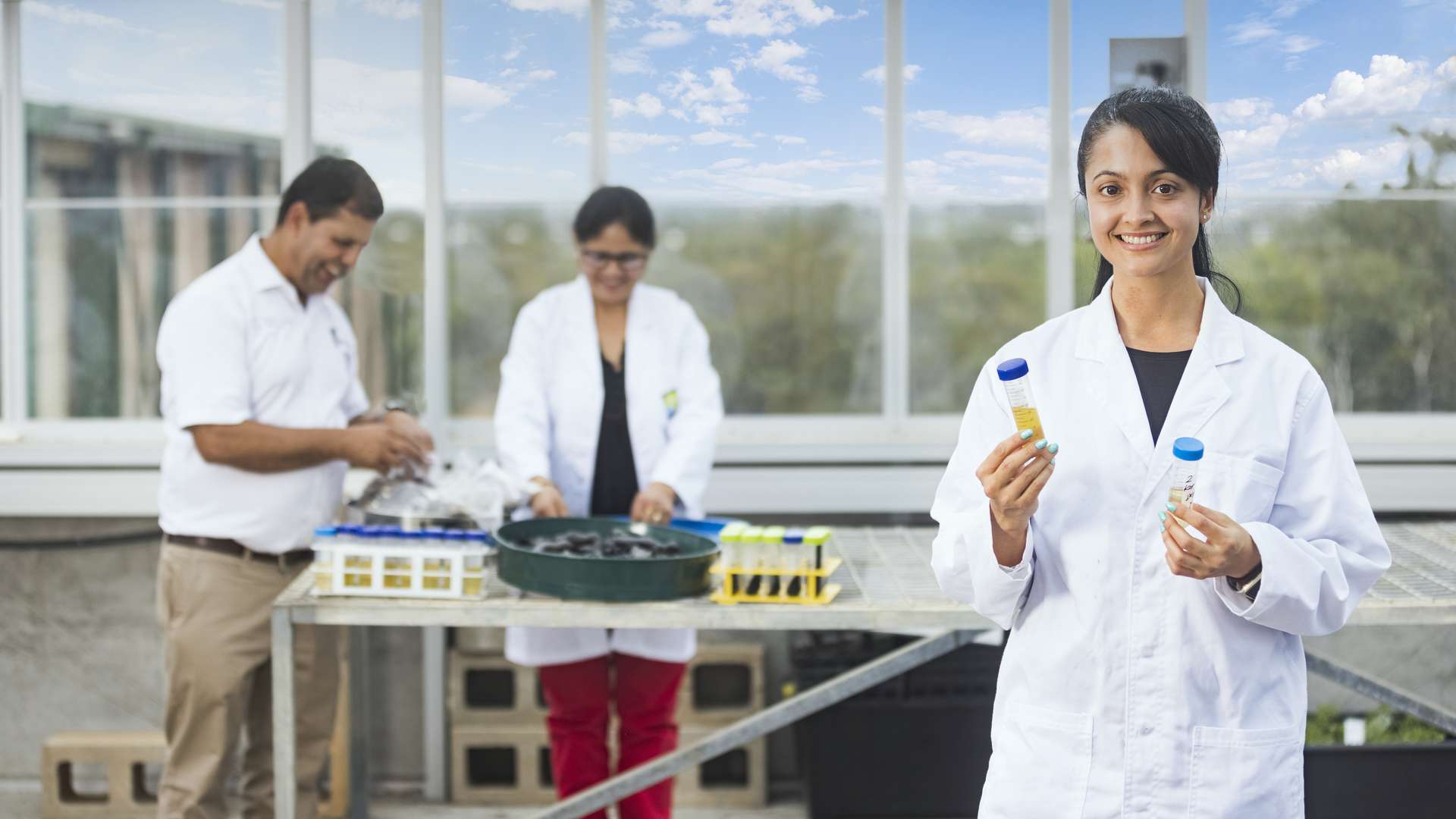 A research student in a lab coat stands holding samples while other researchers examine plant samples in the background.