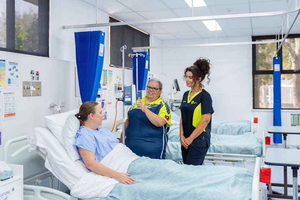 A nursing lecturer assists a nursing student in caring for a patient who is in bed in a realistic hospital setting.