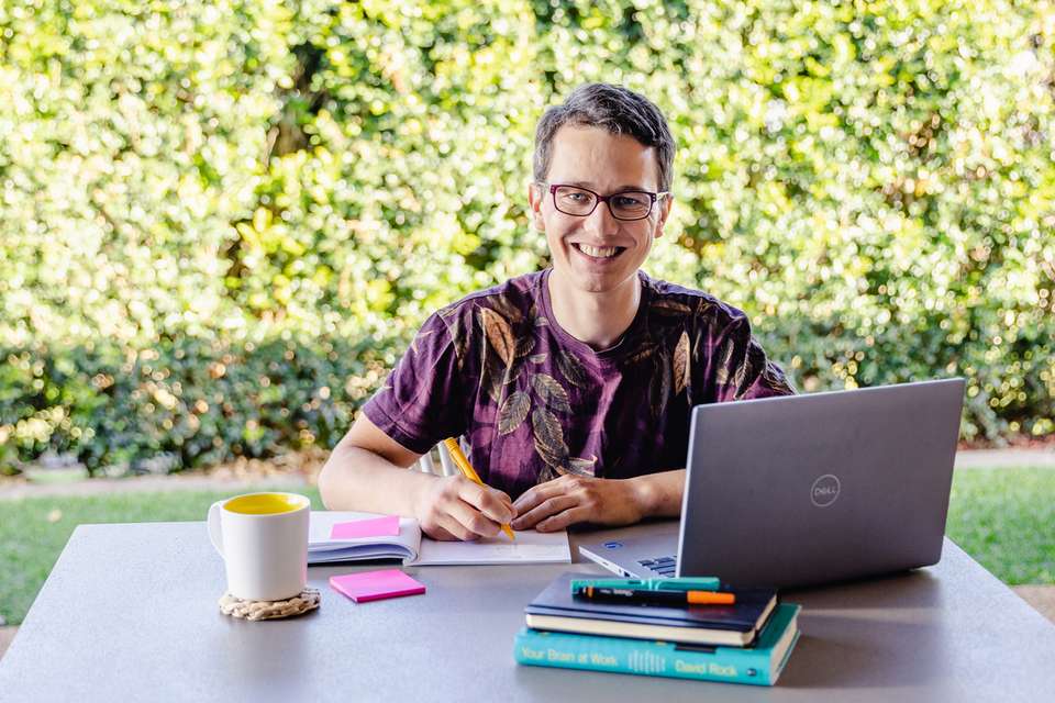 Student smiling while studying outside at table with laptop with green shrubs in the background