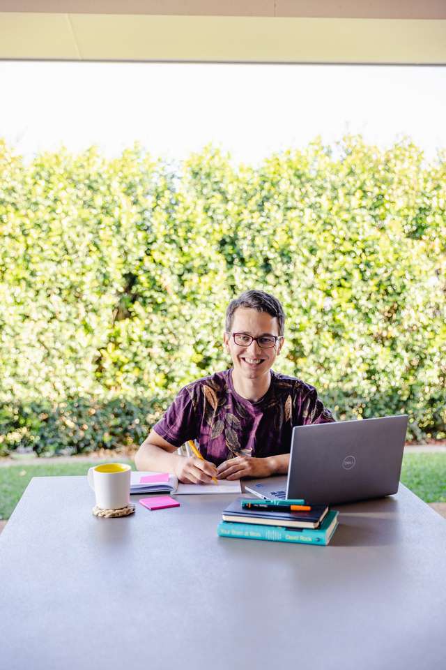 Student smiling while studying outside at table with laptop with green shrubs in the background