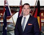 Sean O'Connor smiling at the camera in a suit and tie with three country flags behind